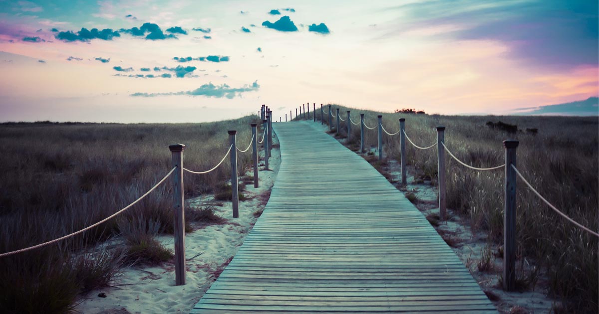 A walkway made of wood extends over a sandy, grassy hill in to the sunset.