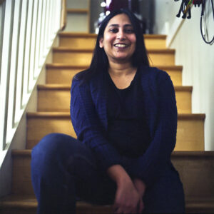 Soumya Natarajan sits on her stairs, smiling, wearing a cardigan and quarter inch cables hang on the wall beside her.
