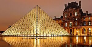 The Louvre in Paris at night. It is a rectangular stone building, and there are warm lights in the windows. A glass pyramid sits in front of it glowing with light and reflecting in the water of a large fountain.