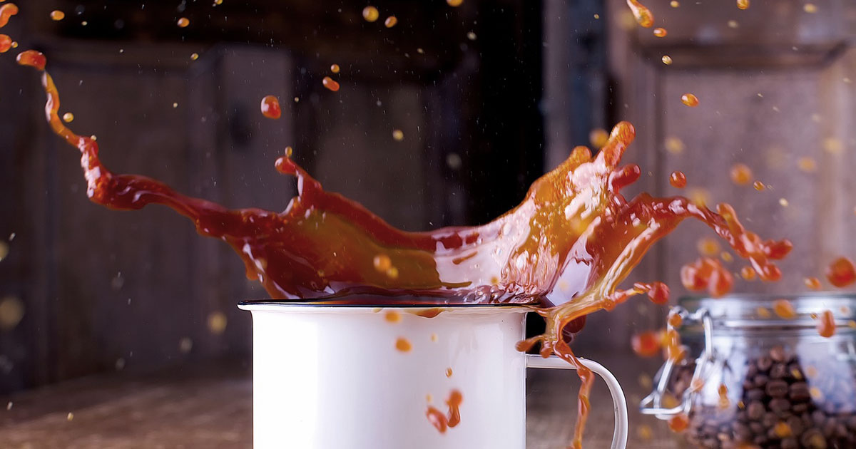 A coffee cup full of coffee is mid explosion and the coffee is flying everywhere across a table.